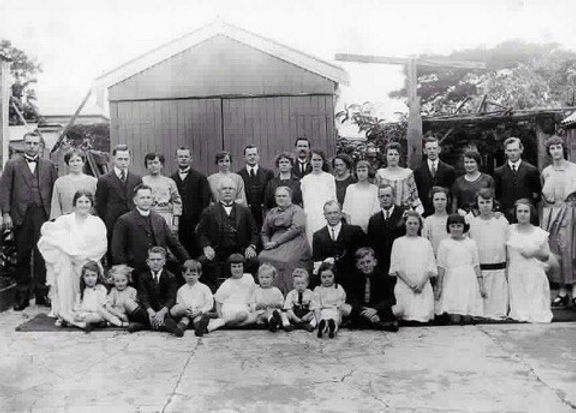 A large group of men, women, and children pose for a black-and-white photo in front of a wooden shed—an early glimpse into H Parsons history, celebrating 130 years as a trusted local funeral service. by H Parsons Funeral Directors