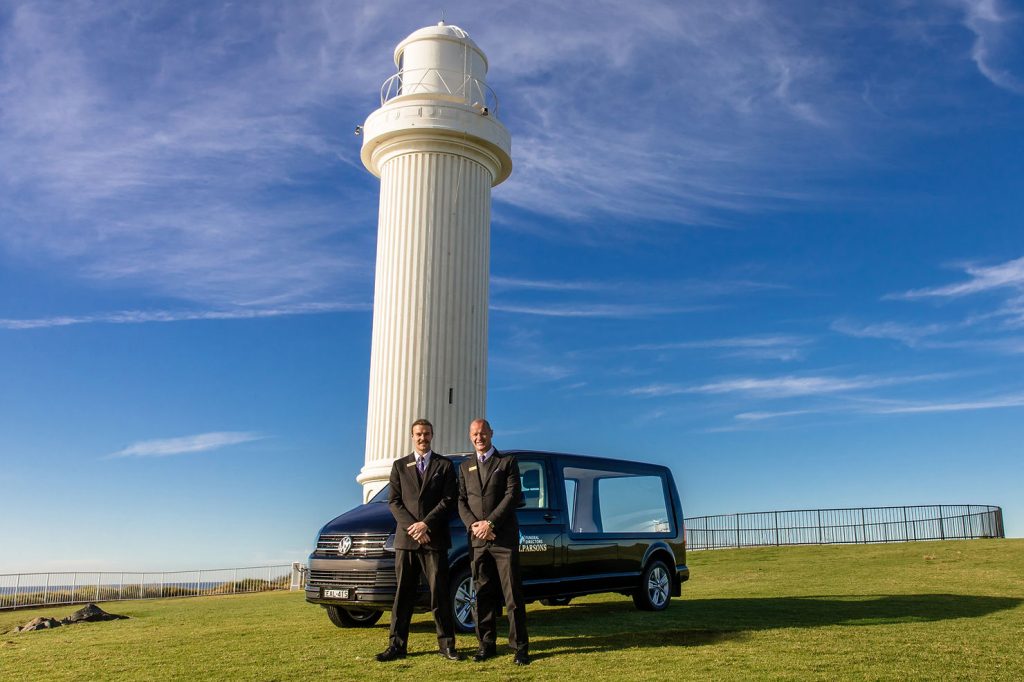 Two men in dark suits stand in front of a black hearse, representing H Parsons History as renowned Wollongong funeral directors, near a tall white lighthouse under a blue sky with wispy clouds. by H Parsons Funeral Directors
