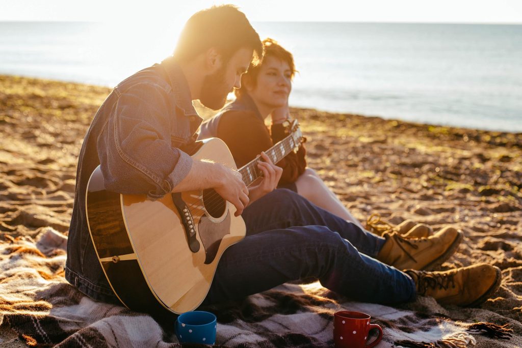 Two people sit on a blanket at the beach during sunset. One plays an acoustic guitar, quietly strumming best funeral songs as the other listens. Two mugs sit in front of them, with the sea stretching out in the background. by H Parsons Funeral Directors