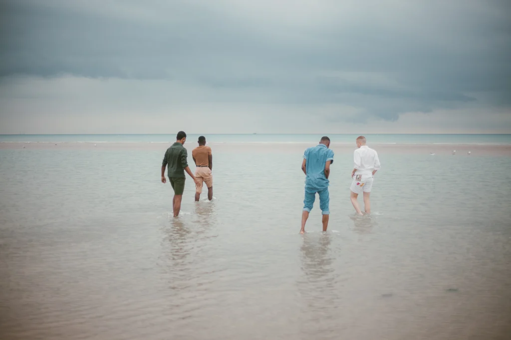 Four people walk away from the camera through shallow water at a beach under a cloudy sky, each wearing differently coloured clothing. The calm and peaceful scene evokes reflection, much like when a funeral applicant considers who is responsible for arranging funeral plans. by H Parsons Funeral Directors