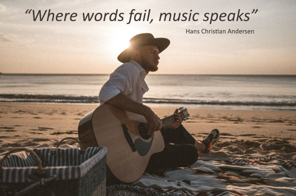 A man in a hat sits on a blanket at the beach during sunset, playing an acoustic guitar. Text above reads, “Where words fail, music speaks” – Hans Christian Andersen—a reminder of how uplifting funeral songs can touch our hearts. by H Parsons Funeral Directors