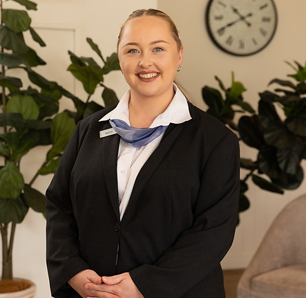 A woman wearing a black blazer, white shirt, and a name badge stands smiling indoors. Behind her are green plants, a clock on the wall, and part of a beige chair. by H Parsons Funeral Directors