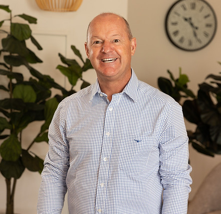 A smiling, bald man in a checked button-up shirt stands indoors in front of large leafy plants and a round wall clock. by H Parsons Funeral Directors