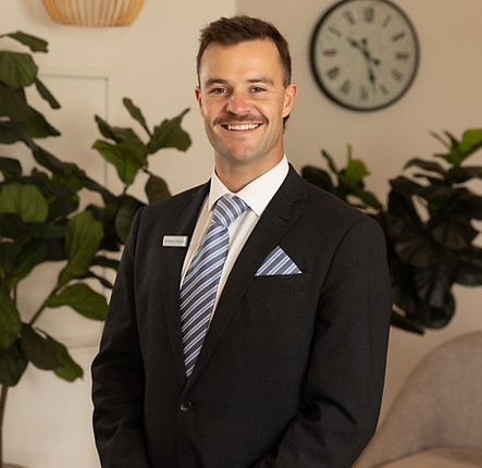 A man in a dark suit and striped tie stands smiling indoors. He has short hair and a moustache. Behind him are green plants, a wall clock, and neutral-coloured decor. by H Parsons Funeral Directors