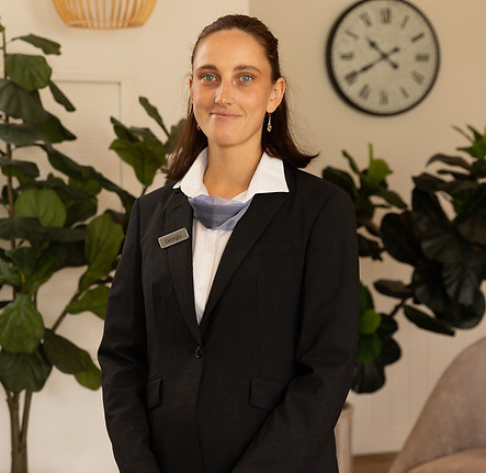 A woman in a dark suit jacket and white shirt stands indoors, smiling slightly. She wears a name badge and a scarf at her collar. Behind her are green plants and a wall clock showing around 10:10. by H Parsons Funeral Directors