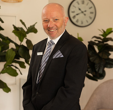 A smiling man in a black suit and striped tie stands indoors with green plants and a wall clock in the background. He wears a name badge and has a neatly trimmed beard and short hair. by H Parsons Funeral Directors