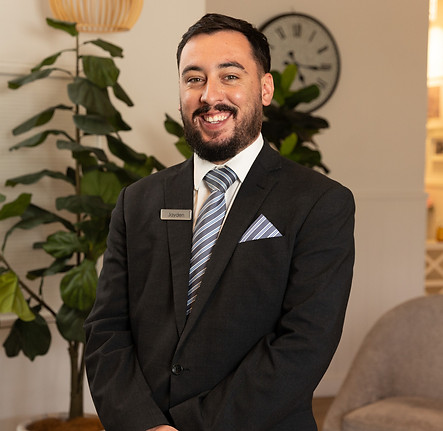 A man in a dark suit, striped tie, and name badge smiles whilst standing indoors near a leafy plant and a wall clock. The background includes home décor elements and warm lighting. by H Parsons Funeral Directors