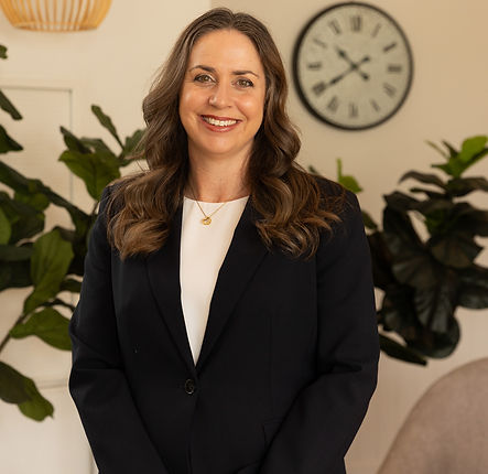 A woman with long brown hair wearing a black blazer and white shirt smiles at the camera. She stands indoors in front of green plants and a wall clock showing 10:09. by H Parsons Funeral Directors