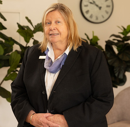 A woman with blonde hair wearing a black blazer, white shirt, and blue scarf stands indoors in front of green plants and a wall clock, smiling gently at the camera. by H Parsons Funeral Directors