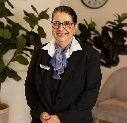 A woman wearing glasses, a dark suit, and a blue scarf stands smiling indoors. She has a name badge and is positioned in front of large green plants and a beige chair. by H Parsons Funeral Directors