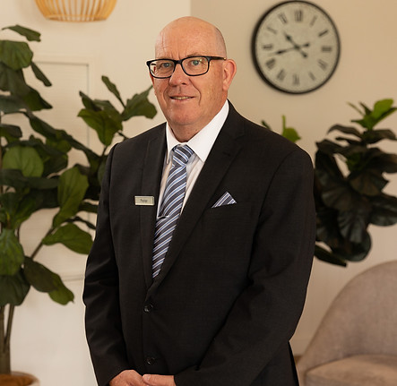 A man in a dark suit, striped tie, and glasses stands indoors, smiling slightly. He has a name badge on his jacket. Behind him are green plants, a grey chair, and a wall clock showing 10:10. by H Parsons Funeral Directors