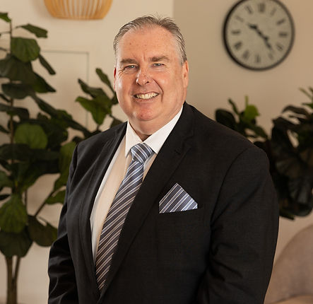 A middle-aged man in a dark suit, white shirt, and striped tie smiles at the camera. He stands indoors near leafy green plants, with a clock and decorative items on the wall in the background. by H Parsons Funeral Directors