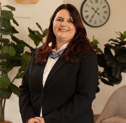 A woman with long brown hair, wearing a black blazer and white shirt, stands smiling in a modern office setting with green plants and a large wall clock in the background. by H Parsons Funeral Directors