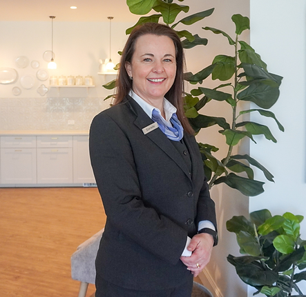 A woman in a dark business suit stands smiling indoors beside large green plants. The background features a bright, modern kitchen with pendant lights, white cupboards, and decorative wall plates. by H Parsons Funeral Directors
