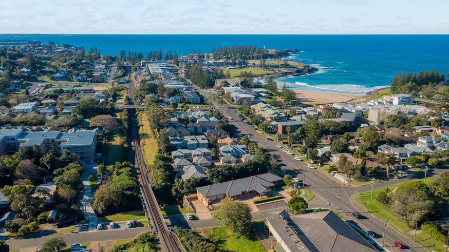 Aerial view of a coastal town with houses, roads, greenery, the Kiama Funeral Chapel, a railway line, and a beach meeting the blue sea under a partly cloudy sky. by H Parsons Funeral Directors