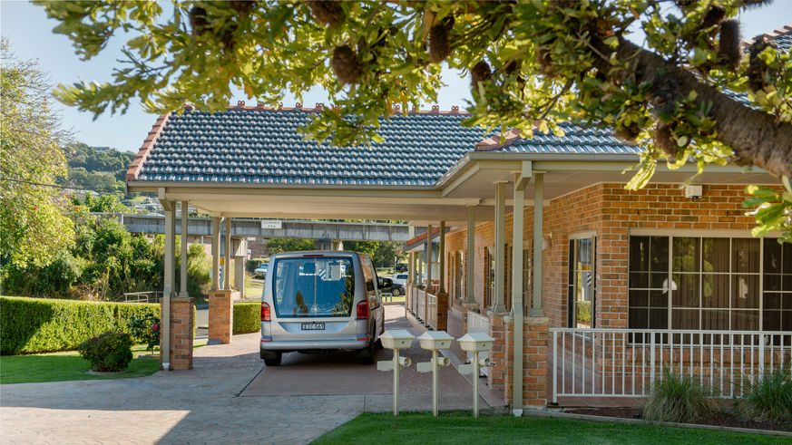 A silver van is parked in the carport of a single-storey brick house with a tiled roof, possibly near Kiama Funeral Chapel. The house has a manicured lawn, a tree in the foreground, and several postboxes by the driveway. by H Parsons Funeral Directors