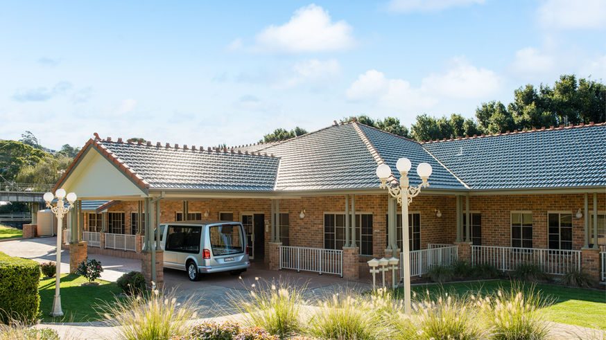 A large, single-storey brick house with a grey tiled roof sits amid green lawns and landscaped bushes, serving as the Kiama Funeral Chapel. A silver van is parked under a covered drive near the entrance, ready for funeral services in Kiama. by H Parsons Funeral Directors
