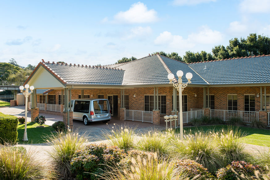 A large, single-storey brick house with a grey tiled roof sits amid green lawns and landscaped bushes, serving as the Kiama Funeral Chapel. A silver van is parked under a covered drive near the entrance, ready for funeral services in Kiama. by H Parsons Funeral Directors
