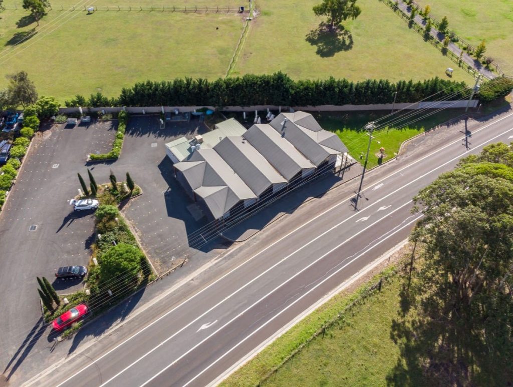 Aerial view of a single-storey building with a dark roof and adjacent car park, possibly linked to Shoalhaven Funerals. Located beside a two-lane road, it’s surrounded by grassy fields and trees, with several cars parked near the building. by H Parsons Funeral Directors