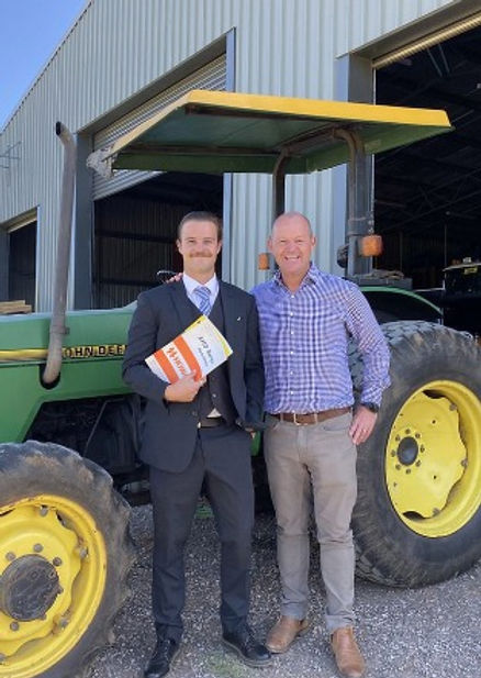 Two men stand smiling in front of a green tractor outside a large metal building. One man wears a suit and holds paperwork, possibly discussing the H Parsons acquisition; the other is casually dressed in a checked shirt and chinos. by H Parsons Funeral Directors