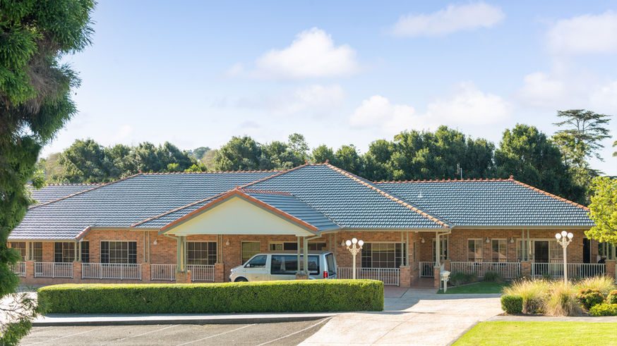 A single-storey brick building with a grey tiled roof, surrounded by greenery and trees, features a covered entrance at Kiama Funeral Chapel, with a van parked nearby and an adjacent car park in the foreground. by H Parsons Funeral Directors