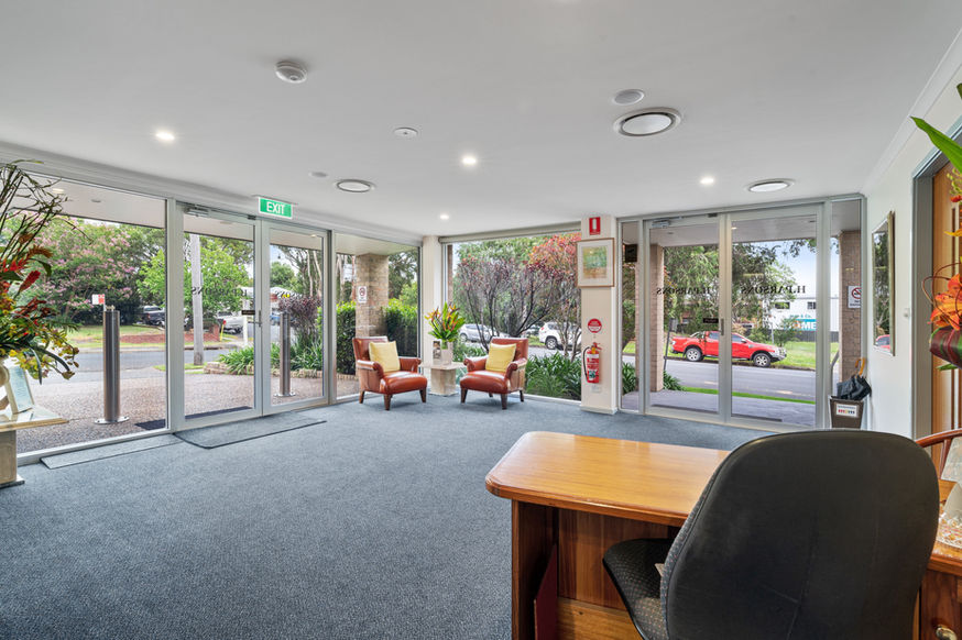 A bright, modern lobby at Nowra Funeral Chapel features large windows, two armchairs, indoor plants, and a wooden reception desk. Glass doors reveal a street with parked cars and greenery outside. by H Parsons Funeral Directors