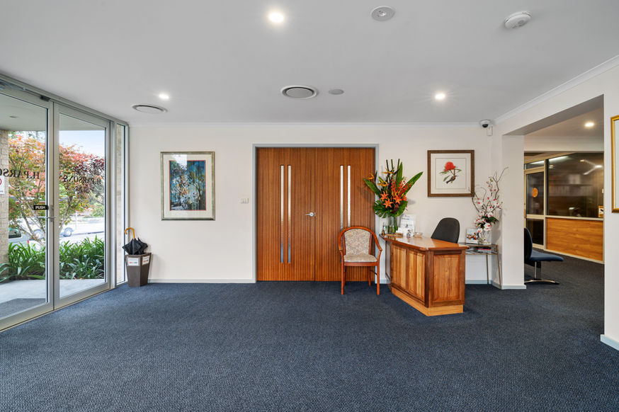 A bright, carpeted reception area at Nowra Funeral Chapel features wooden double doors in the centre, a wooden desk with flowers on the right, a chair, framed artwork on the walls, and glass entrance doors on the left. by H Parsons Funeral Directors