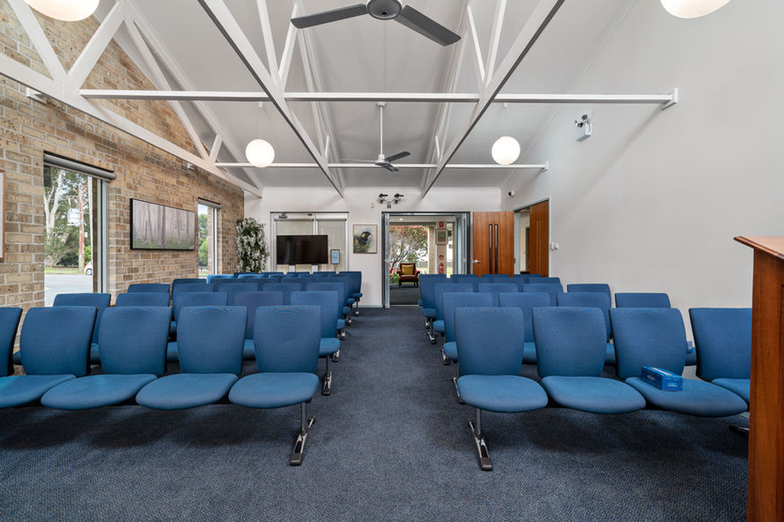 Rows of blue chairs are arranged in a bright, modern room with high ceilings, exposed beams, and large windows at Nowra Funeral Chapel. A lectern stands to the right, with a television and paintings adorning the walls. by H Parsons Funeral Directors