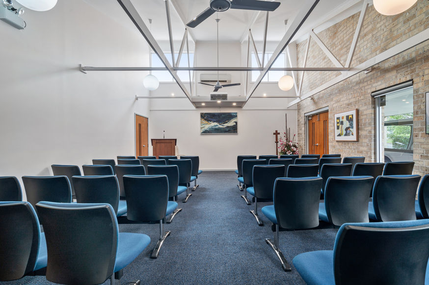 Rows of blue chairs face a lectern in a bright, modern room at Nowra Funeral Chapel, with exposed brick walls, large windows, and hanging lights. A painting and floral arrangement decorate the front wall, creating a serene setting for funeral services. by H Parsons Funeral Directors