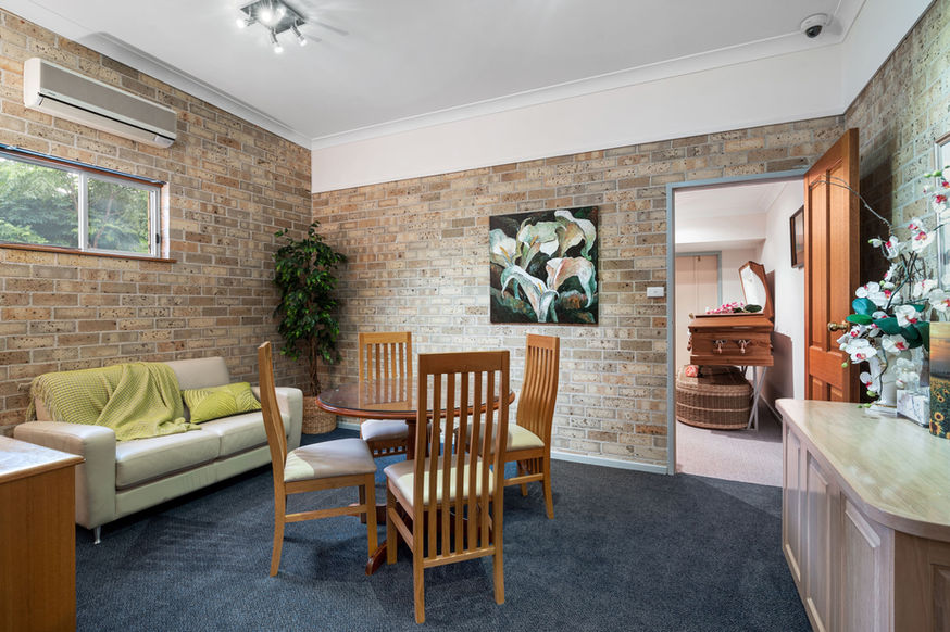 A cosy living-dining area with exposed brick walls, a small sofa, round table with four wooden chairs, plants, and a wall painting welcomes you—just steps from the Nowra Funeral Chapel—through an open door leading to a room with a chest of drawers and laundry basket. by H Parsons Funeral Directors