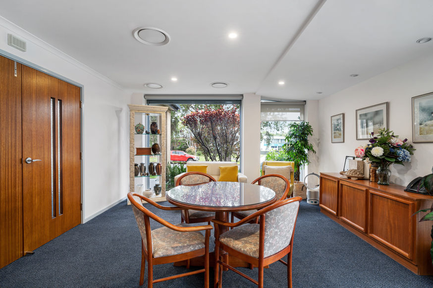 A dining room with a round table and four chairs, glass doors opening to a garden, wooden cupboards, decorative shelves, and framed artwork on white walls. Natural light fills the room, offering a serene view reminiscent of Nowra Funeral Chapel’s peaceful surroundings. by H Parsons Funeral Directors