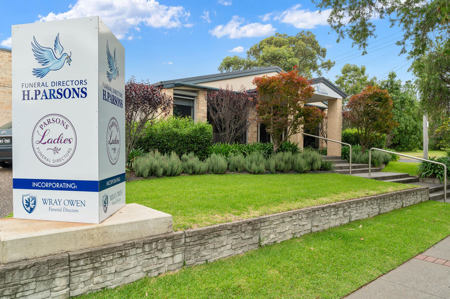 A funeral home building, known as Nowra Funeral Chapel, with a large sign reading "Funeral Directors H. Parsons" and "Parsons Ladies," surrounded by green grass, shrubs, and trees on a sunny day. by H Parsons Funeral Directors