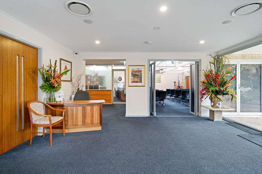 A bright, modern office reception area at Nowra Funeral Chapel features a wooden desk, a chair, floral arrangements, framed art, and glass doors leading to a meeting room with chairs. The floor is carpeted in blue. by H Parsons Funeral Directors