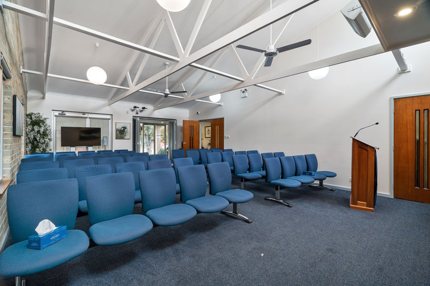 Rows of blue padded chairs are arranged facing a wooden lectern in the bright, modern Nowra Funeral Chapel, featuring white walls, high ceiling, fans, and large windows—an elegant setting for funeral services in Nowra. by H Parsons Funeral Directors