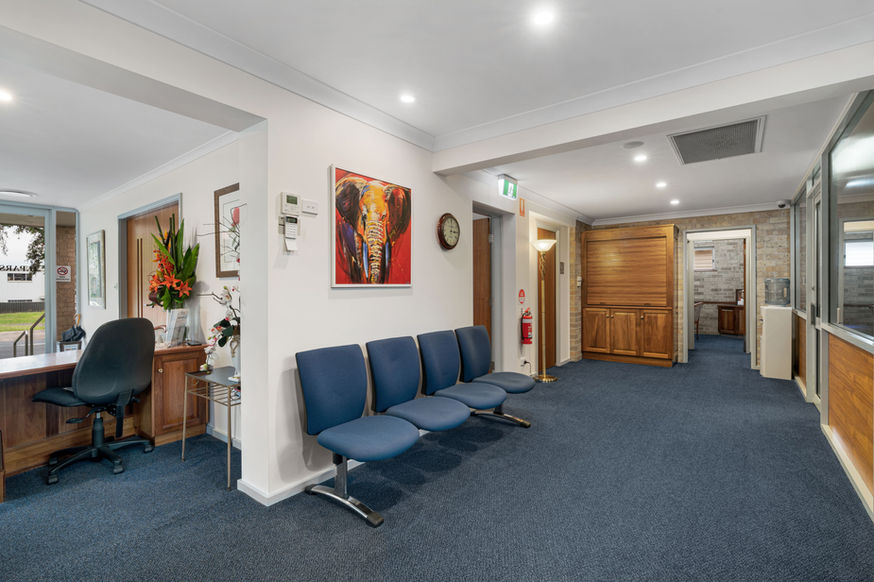 A clean, well-lit waiting area at Nowra Funeral Chapel features blue chairs, a reception desk with flowers, an elephant painting on the wall, and doors to other rooms. The space is finished with blue carpet and white walls. by H Parsons Funeral Directors