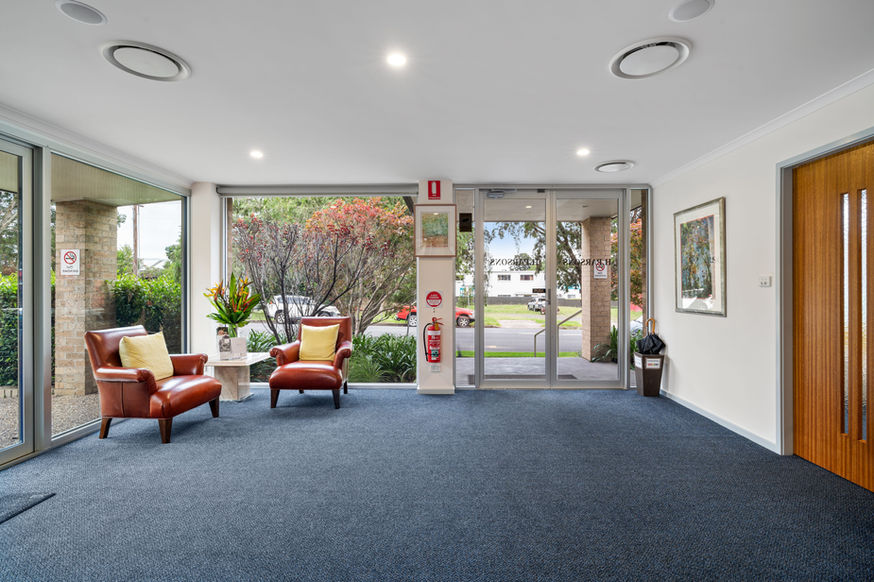 A bright lobby at Nowra Funeral Chapel features two brown leather chairs with yellow cushions, a small table with plants, large windows, wall art, and peaceful views of greenery and the street outside. by H Parsons Funeral Directors