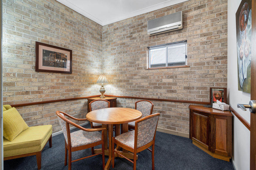 A small room with exposed brick walls, a round wooden table with four chairs, a yellow cushioned bench, a lamp, air conditioner, two framed pictures, and a corner cupboard under a window at Nowra Funeral Chapel. by H Parsons Funeral Directors