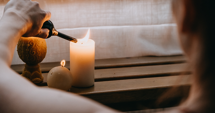 A person uses a lighter to ignite a white pillar candle on a wooden surface, with a round candle and decorative object nearby—an elegant setting that inspires memorial ideas as light filters through a sheer curtain in the background. by H Parsons Funeral Directors
