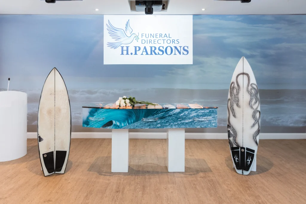 A surfboard serves as a coffin, adorned with white flowers, and stands between two upright surfboards. A sign above reads "Funeral Directors H. Parsons"—a nod to eco-friendly burials and emerging funeral trends Australia embraces by the ocean. by H Parsons Funeral Directors