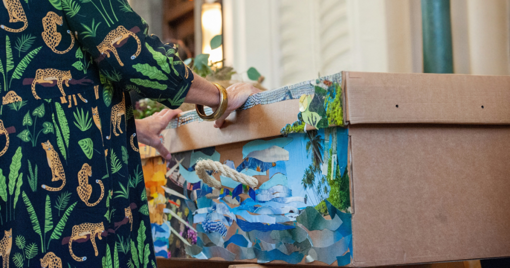A person wearing a green dress with a cheetah and leaf pattern places their hands on a colourfully decorated biodegradable urn, covered in nature-themed collage art, reflecting the spirit of green burial Australia. by H Parsons Funeral Directors