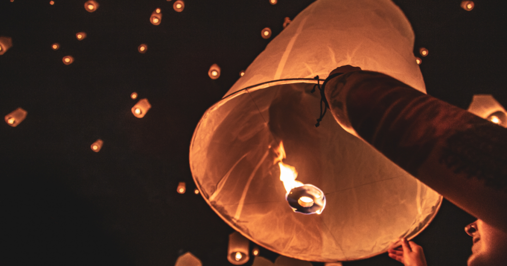 A person releases a glowing paper lantern into the night sky, joining many others floating upwards—a beautiful tribute and one of many unique celebration of life ideas, with warm candlelight illuminating the scene against the dark background. by H Parsons Funeral Directors