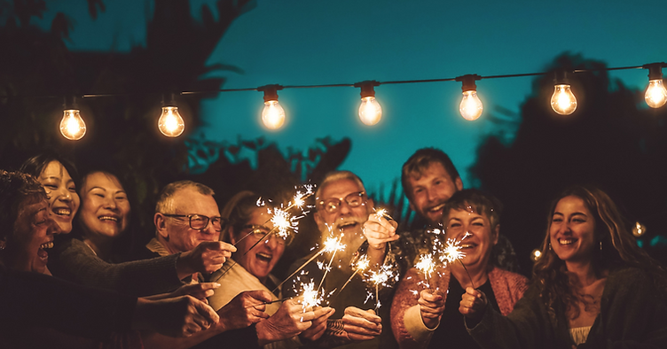 A group of people smiling and holding sparklers together at night under fairy lights, sharing joyful moments outdoors with trees in the background—perfect inspiration for life celebration ideas. by H Parsons Funeral Directors
