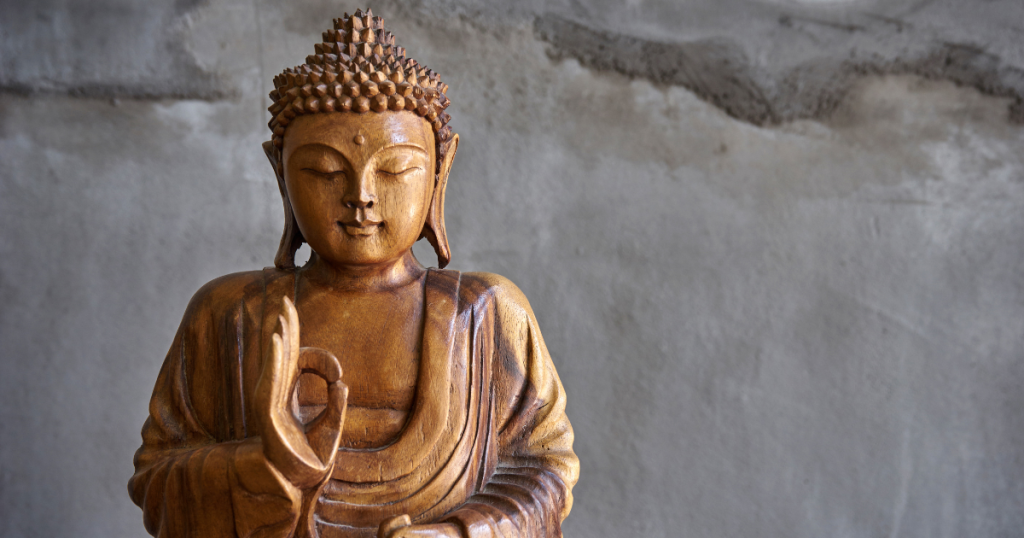 A wooden Buddha statue with one hand raised in blessing or teaching, set against a plain, textured grey wall—echoing serenity found in Buddhist funeral traditions. by H Parsons Funeral Directors