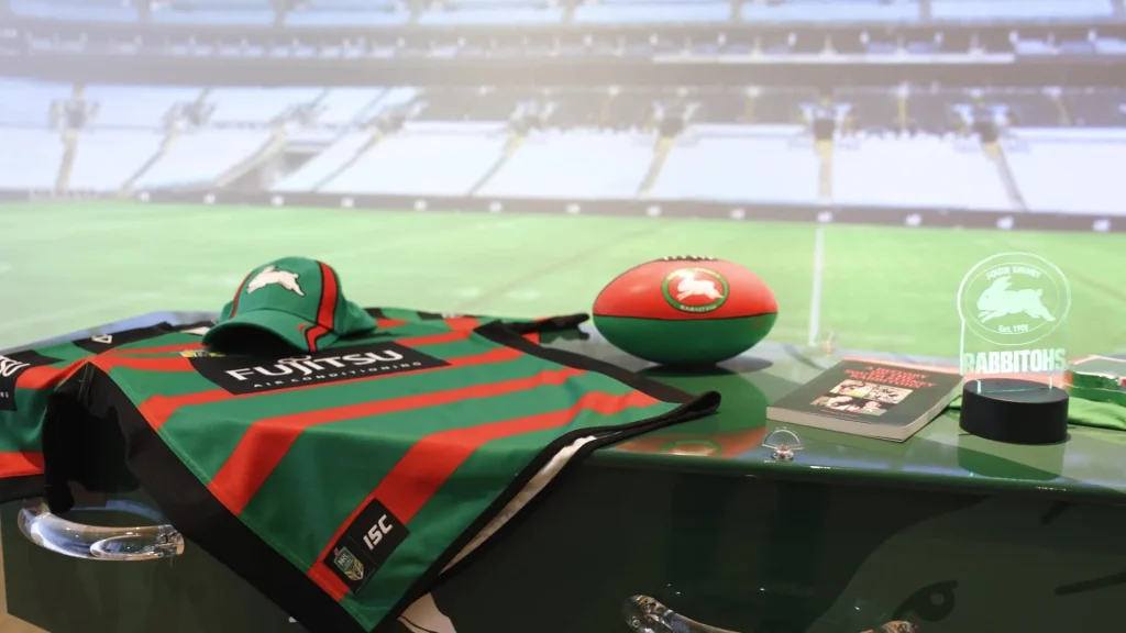 A green, red, and black South Sydney Rabbitohs rugby jersey, cap, and ball are displayed on a counter in front of a blurred stadium backdrop, alongside a booklet and wristband—an unexpected nod to funeral trends Australia now embraces. by H Parsons Funeral Directors