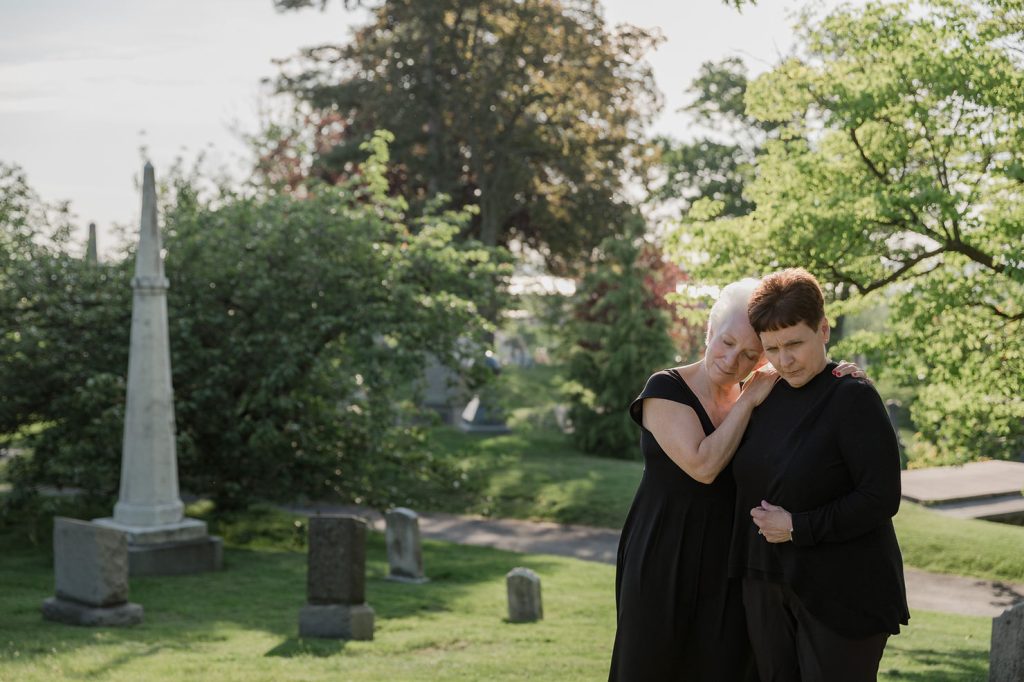 Two women dressed in black funeral clothing comfort each other in a cemetery on a sunny day, surrounded by gravestones and green trees. One woman rests her head on the other's shoulder. by H Parsons Funeral Directors