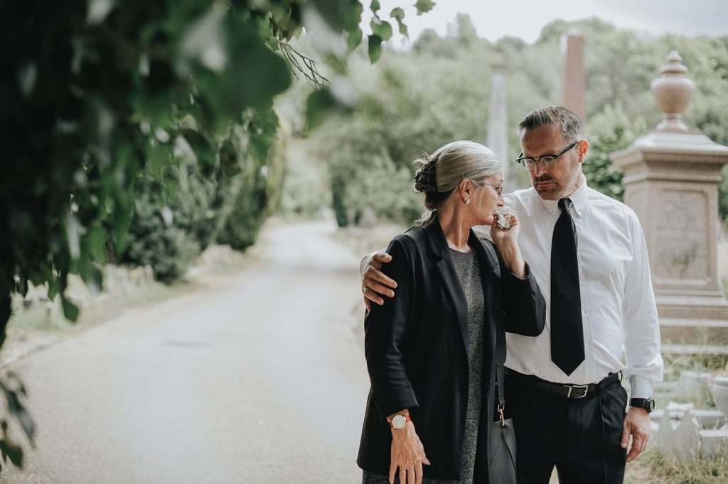 A man and woman dressed in classic funeral attire stand close together in a cemetery, comforting each other. The man has his arm around the woman as she wipes her eye, both appearing sombre. Trees and gravestones are visible in the background. by H Parsons Funeral Directors