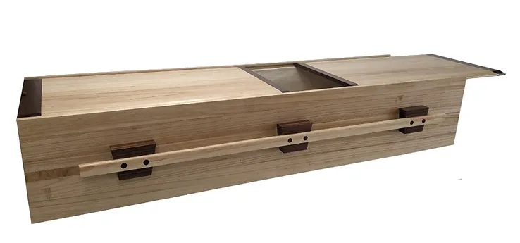 A light wood rectangular coffin with dark wood accents, sliding lid, and two side handles, ideal for green burial, shown against a white background. by H Parsons Funeral Directors