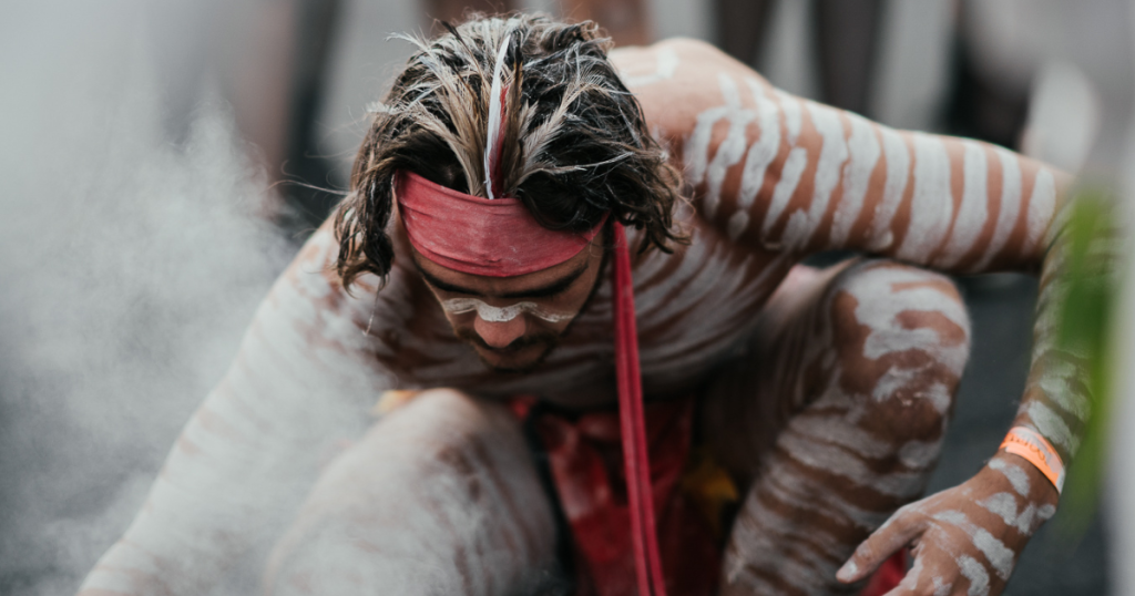 A person with long hair and a red headband is covered in white body paint in a striped pattern, crouching close to the ground and surrounded by white dust during Australian Aboriginal ceremonies. by H Parsons Funeral Directors