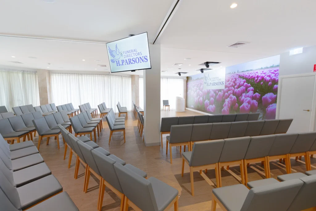 A modern, bright funeral chapel with rows of grey cushioned chairs, a large screen displaying "Funeral Directors PARSONS," and a mural of pink tulips. Natural light fills the room, reflecting new funeral trends Australia like eco-friendly burials. by H Parsons Funeral Directors