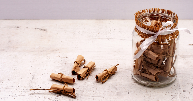 A glass jar decorated with hessian and a white ribbon holds several rolled-up brown paper notes tied with string—an inspiring choice for those seeking unique celebration of life ideas; similar notes are scattered nearby on a white wooden surface. by H Parsons Funeral Directors